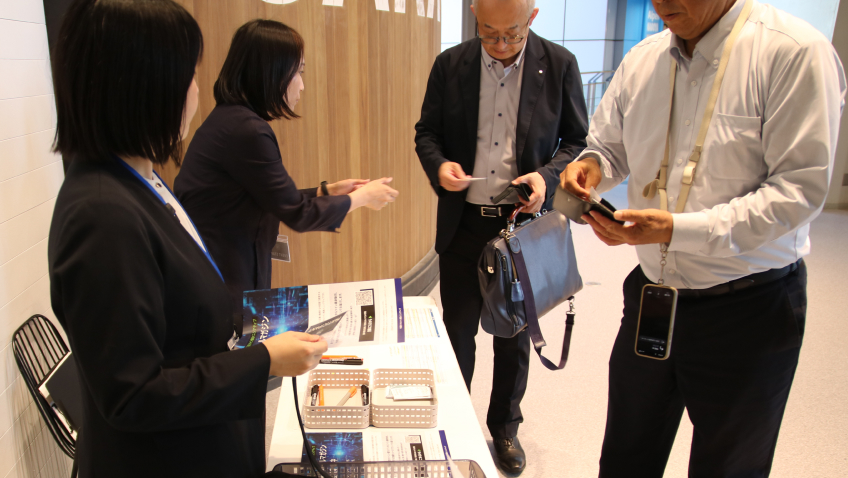 受付の様子。二人の参加者が名刺を提出し、事務局スタッフがそれを受け取り、フライヤーを渡している